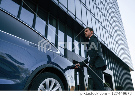 Man in suit and tie is holding charger and standing near his electric car outdoors 127449673