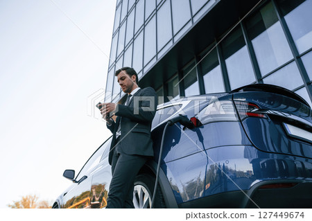 Electric car is charging, businessman with smartphone is waiting Electric car is charging, businessman with smartphone is waiting 127449674