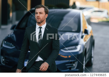 Standing, in black suit. Businessman is near his car outdoors Standing, in black suit. Businessman is near his car outdoors 127449685