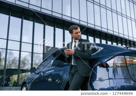 Electric car is charging, businessman with smartphone is waiting Electric car is charging, businessman with smartphone is waiting 127449694