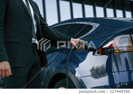 Close up view, putting charger into the socket. Businessman is standing near his electric car outdoors Close up view, putting charger into the socket. Businessman is standing near his electric car outdoors 127449700