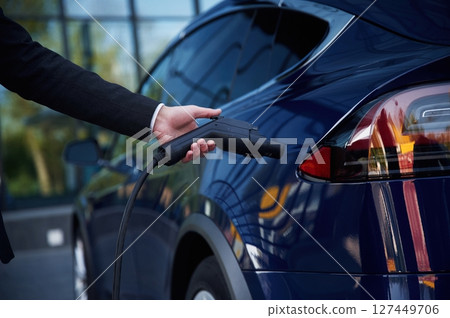 Hand is holding charger, close up view. Businessman is standing near his electric car outdoors Hand is holding charger, close up view. Businessman is standing near his electric car outdoors 127449706