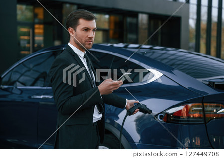 Man in suit and tie is holding charger and standing near his electric car outdoors Man in suit and tie is holding charger and standing near his electric car outdoors 127449708