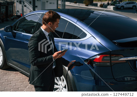 Businessman is standing near his electric car outdoors. Using smartphone Businessman is standing near his electric car outdoors. Using smartphone 127449712
