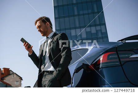 Businessman is standing near his electric car outdoors. Using smartphone Businessman is standing near his electric car outdoors. Using smartphone 127449718