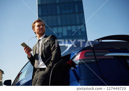 Businessman is standing near his electric car outdoors. Using smartphone Businessman is standing near his electric car outdoors. Using smartphone 127449720