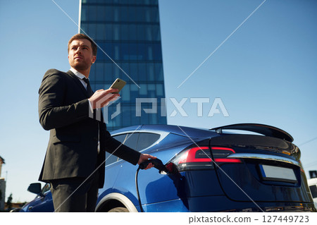 Businessman is standing near his electric car outdoors. Using smartphone 127449723