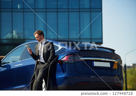 Business building behind. Man in suit is standing near his electric car outdoors Business building behind. Man in suit is standing near his electric car outdoors 127449728