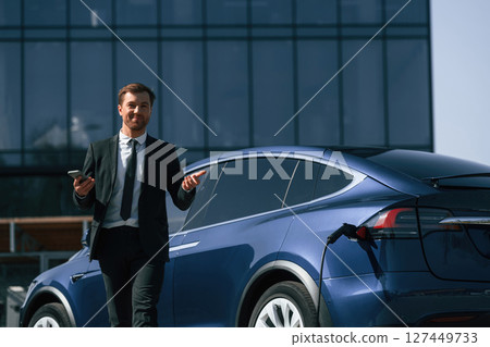 Business building behind. Man in suit is standing near his electric car outdoors Business building behind. Man in suit is standing near his electric car outdoors 127449733
