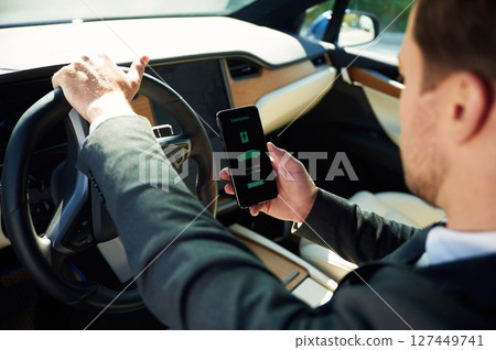 Holding smartphone with charging progress app. Businessman in suit is driving his electric car 127449741