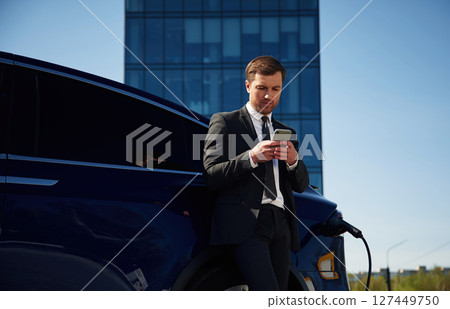 Waiting for a charge. Businessman is standing near his electric car outdoors Waiting for a charge. Businessman is standing near his electric car outdoors 127449750