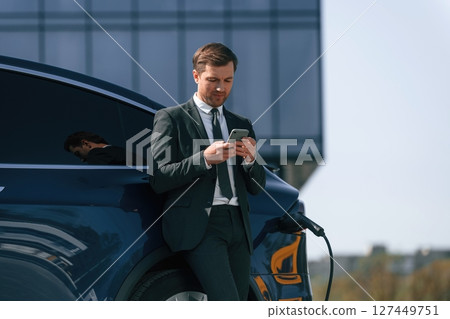 Waiting for a charge. Businessman is standing near his electric car outdoors Waiting for a charge. Businessman is standing near his electric car outdoors 127449751