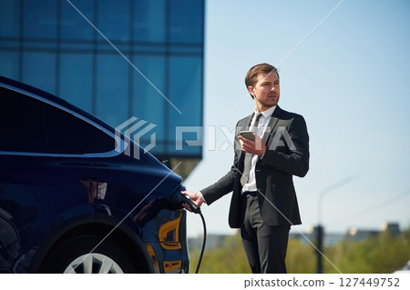 Business building behind. Man in suit is standing near his electric car outdoors 127449752