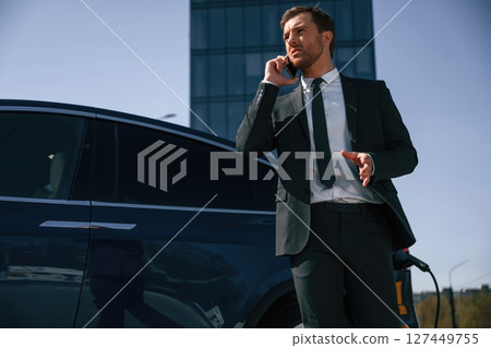 Business call, talking by phone. Man is standing near his electric car outdoors 127449755