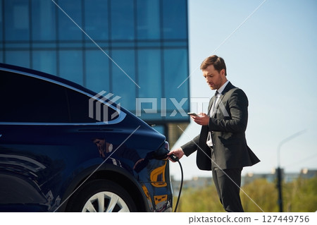 Holding the charger. Businessman is standing near his electric car outdoors 127449756