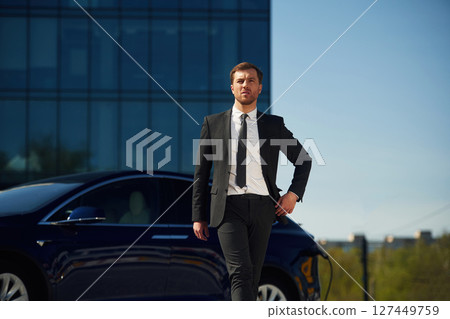Serious facial expression. Businessman is standing near his electric car outdoors Serious facial expression. Businessman is standing near his electric car outdoors 127449759