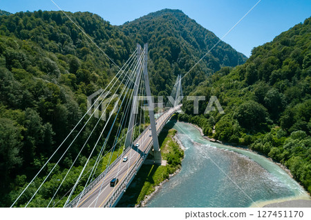 A suspension bridge stretches across a flowing river, surrounded by lush green mountains 127451170