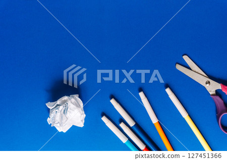 Top view of colorful markers, a crumpled paper ball and scissors arranged on a vivid blue background. School or office supplies creative layout. Top view of colorful markers, a crumpled paper ball and scissors arranged on a vivid blue background. School or office supplies creative layout. 127451366
