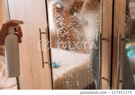 Close-up of a hand spraying glass cleaner on a mirrored wardrobe door during home cleaning. Close-up of a hand spraying glass cleaner on a mirrored wardrobe door during home cleaning. 127451373