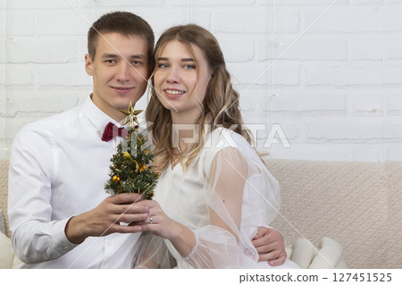 The bride and groom are sitting on the couch and holding a Christmas tree. Christmas wedding. The bride and groom are sitting on the couch and holding a Christmas tree. Christmas wedding. 127451525