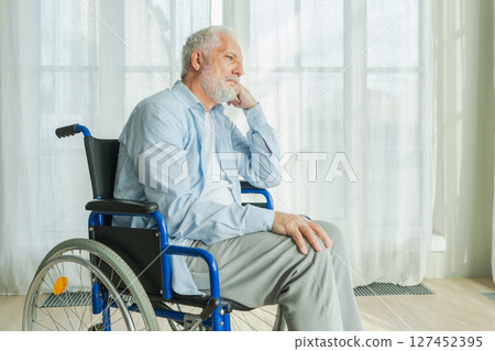 Sad senior man sitting in wheelchair looking out window in retirement home hospital. Old paralyzed man in chair for people with disability feel depressed. Lonely elderly sick man sitting on wheelchair 127452395