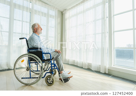 Sad senior man sitting in wheelchair looking out window in retirement home hospital. Old paralyzed man in chair for people with disability feel depressed. Lonely elderly sick man sitting on wheelchair 127452400