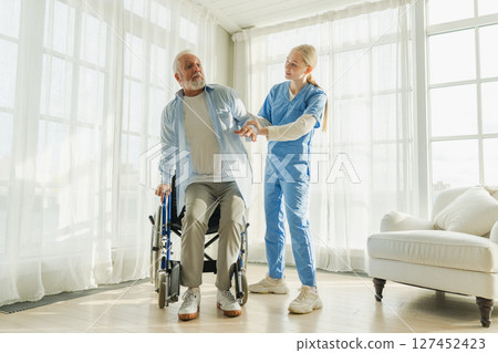 Rehabilitation healthcare. Physiotherapist nurse helps old man patient in wheelchair stand up walking. Man in chair for people with disability doing few steps with nurse helping. Medical caregiver 127452423