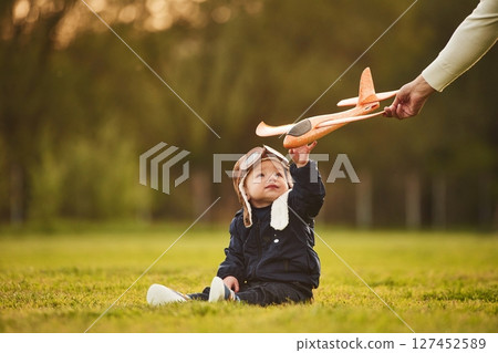 Little boy in aviator glasses is sitting on the field. Woman's hand giving toy plane to him 127452589
