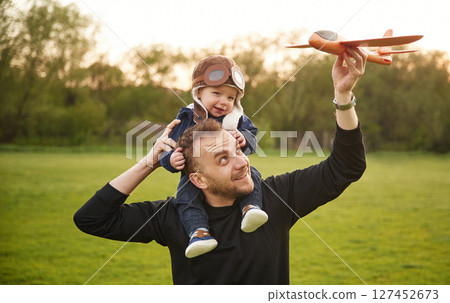 Boy is sitting on man's shoulders. Father with his little son playing with toy plane on the field 127452673