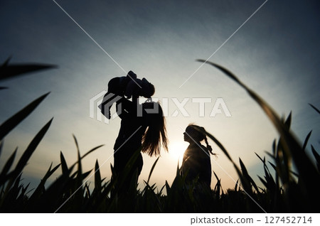 Silhouette of woman that holding her daughter in hands. Another girl is standing and watching Silhouette of woman that holding her daughter in hands. Another girl is standing and watching 127452714