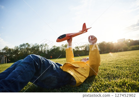 Lying down on ground. Little boy is playing with toy plane on the summer field Lying down on ground. Little boy is playing with toy plane on the summer field 127452902