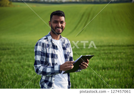 Standing and holding notepad. Handsome Indian man is on the agricultural field 127457061