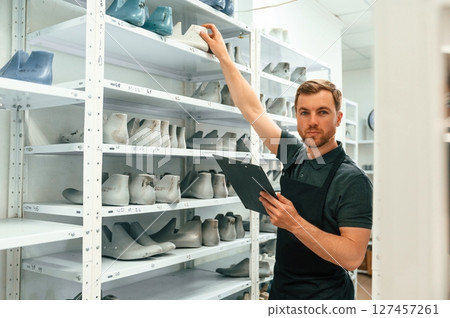 Standing with notepad and near shelves with shoes for prosthesis. Technician working in modern laboratory Standing with notepad and near shelves with shoes for prosthesis. Technician working in modern laboratory 127457261