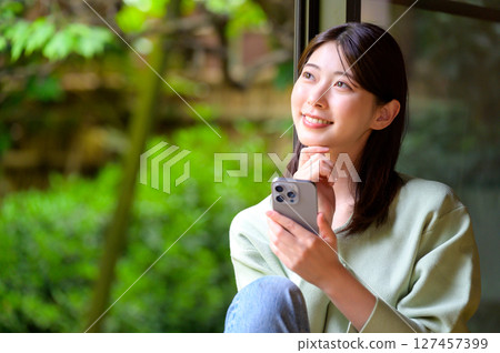 下午，一位年輕女子一邊看著智慧型手機，一邊安詳地微笑，背景是大自然 127457399