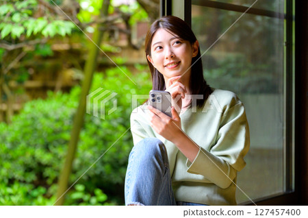 一位年輕女子一手拿著智慧型手機，用溫柔的目光凝視著外面的風景的日常生活場景 127457400