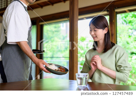 A Japanese dining table where a man wearing an apron serves food with a smile and a woman clasps her hands together to express her gratitude A Japanese dining table where a man wearing an apron serves food with a smile and a woman clasps her hands together to express her gratitude 127457489
