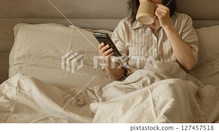 Woman drinking coffee and using phone in bed in the morning. Woman drinking coffee and using phone in bed in the morning. 127457518