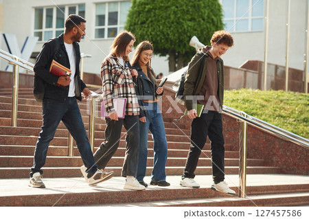 With education material in hands, notepads. Four young students in casual clothes are together outdoors With education material in hands, notepads. Four young students in casual clothes are together outdoors 127457586