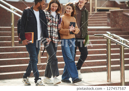 Walking, holding notepads. Four young students in casual clothes are together outdoors 127457587