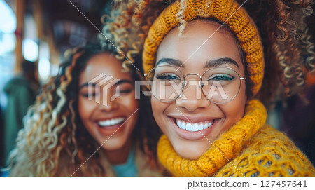Two joyful women with curly hair wearing cozy sweaters and glasses, enjoying a cheerful moment 127457641