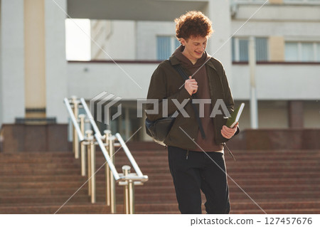 With backpack. Young student with curly hair is outdoors near university 127457676