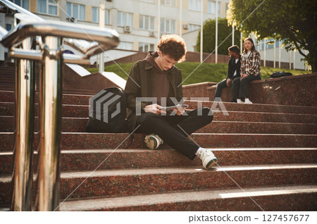 Guy is sitting on the stairs. Young students in casual clothes are together outdoors Guy is sitting on the stairs. Young students in casual clothes are together outdoors 127457677