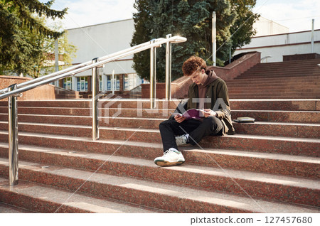 On the stairs, reading. Young student with curly hair is outdoors near university 127457680