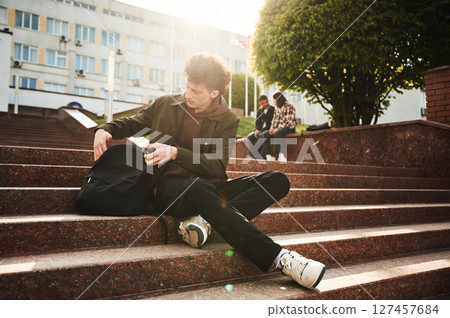 Guy is sitting on the stairs. Young students in casual clothes are together outdoors 127457684
