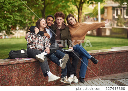 Sitting and smiling. Four young students in casual clothes are together outdoors Sitting and smiling. Four young students in casual clothes are together outdoors 127457868