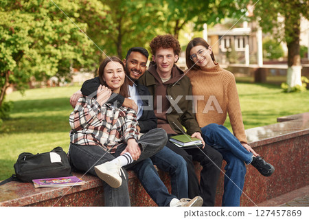 Sitting and smiling. Four young students in casual clothes are together outdoors Sitting and smiling. Four young students in casual clothes are together outdoors 127457869