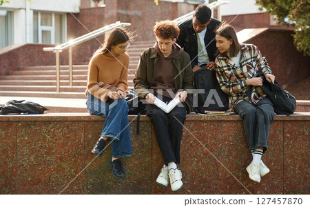 Sitting, reading educational material. Four young students in casual clothes are together outdoors Sitting, reading educational material. Four young students in casual clothes are together outdoors 127457870