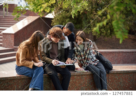 Man is holding notepad, everyone is laughing. Four young students in casual clothes are together outdoors 127457881
