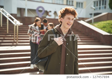 Guy with curly hair is standing in front of his friends. Four young students in casual clothes are together outdoors Guy with curly hair is standing in front of his friends. Four young students in casual clothes are together outdoors 127457894