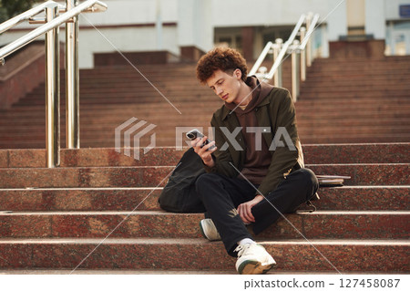 Smartphone is in hands. Young student with curly hair is outdoors near university 127458087